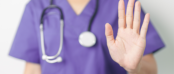 health care worker in scrubs wearing stethoscope with hand held up to signal stop